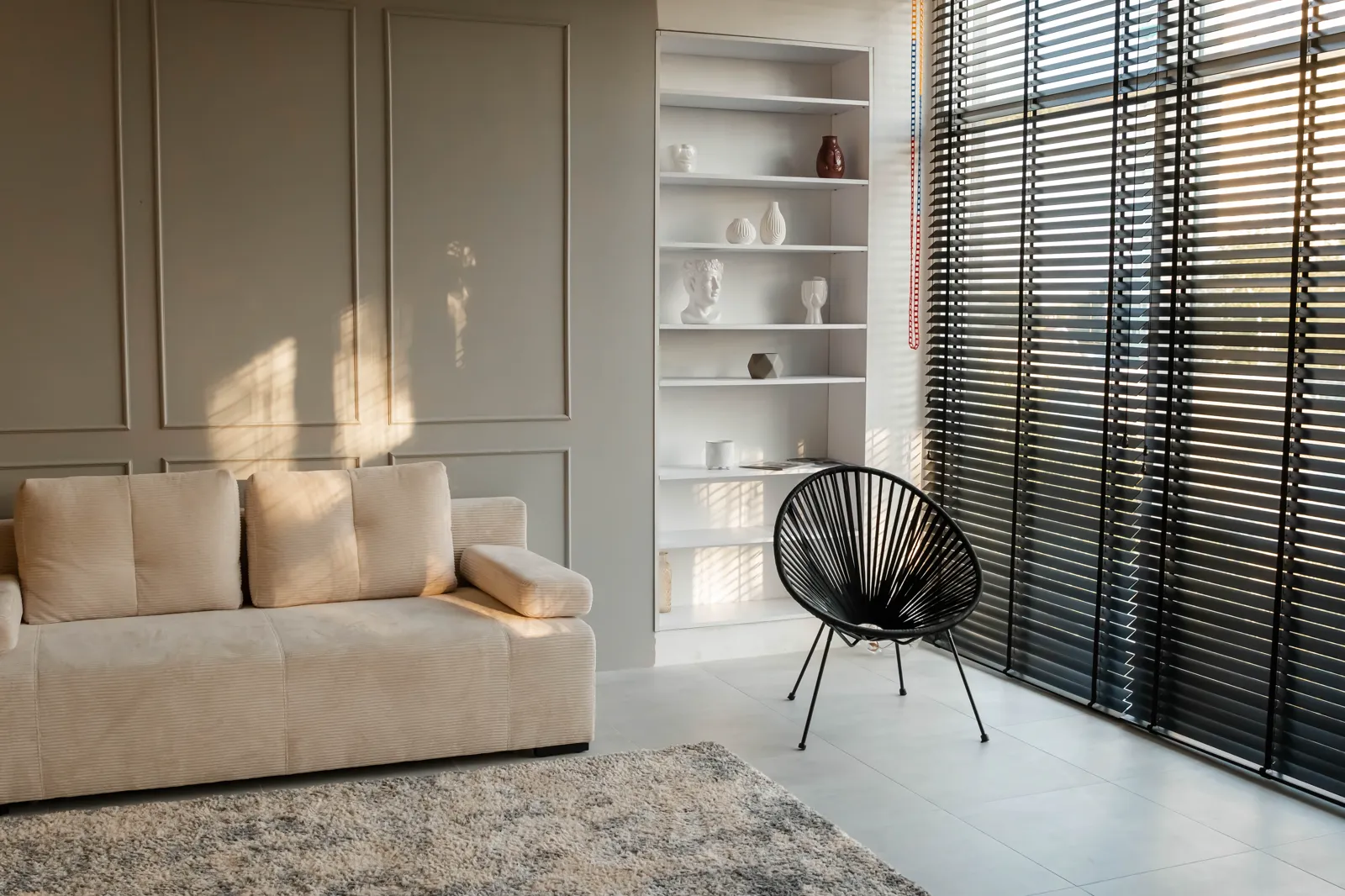 Living room with greige paneled walls, a cream sofa and dark window blinds